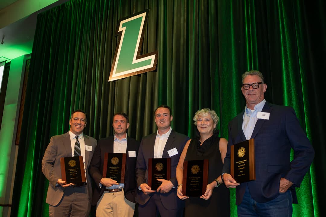 The Hall of Fame Class of 2026 on stage at McGuire Hall, each holding their plaque